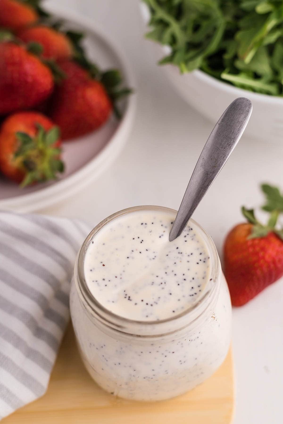 Panera Poppyseed Dressing in jar with spoon, surrounded by strawberries and salad greens. 