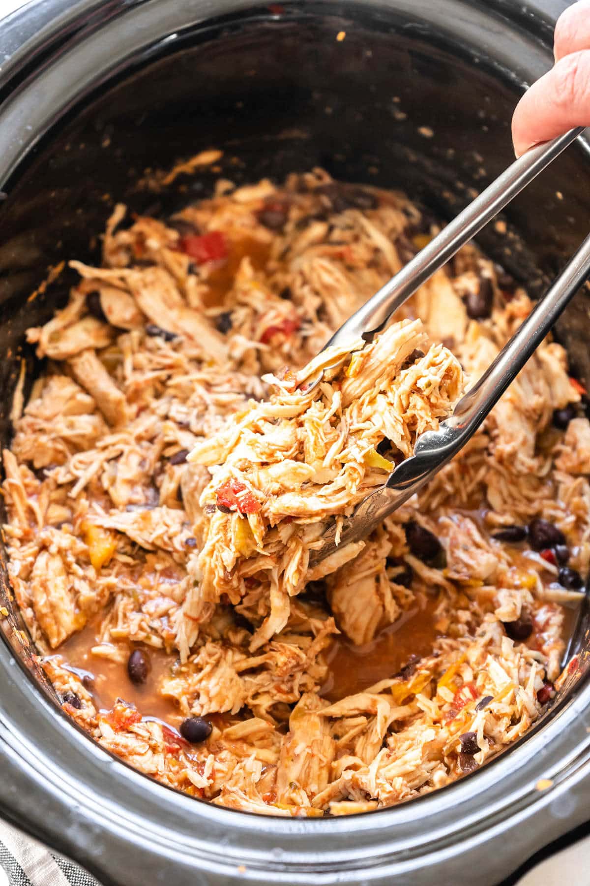 Shredded Chicken taco meat in crockpot with tongs holding juicy chicken.