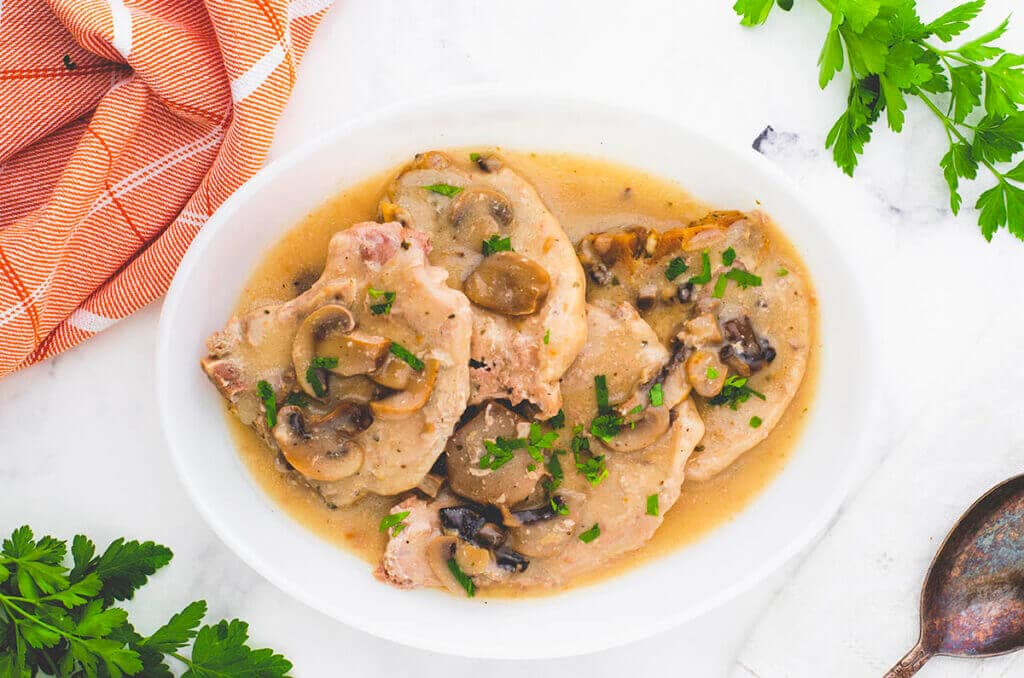 A white oval dish filled with pork chops smothered in a creamy mushroom sauce, garnished with chopped parsley. The dish is surrounded by fresh parsley and an orange checkered cloth on a white marble surface.