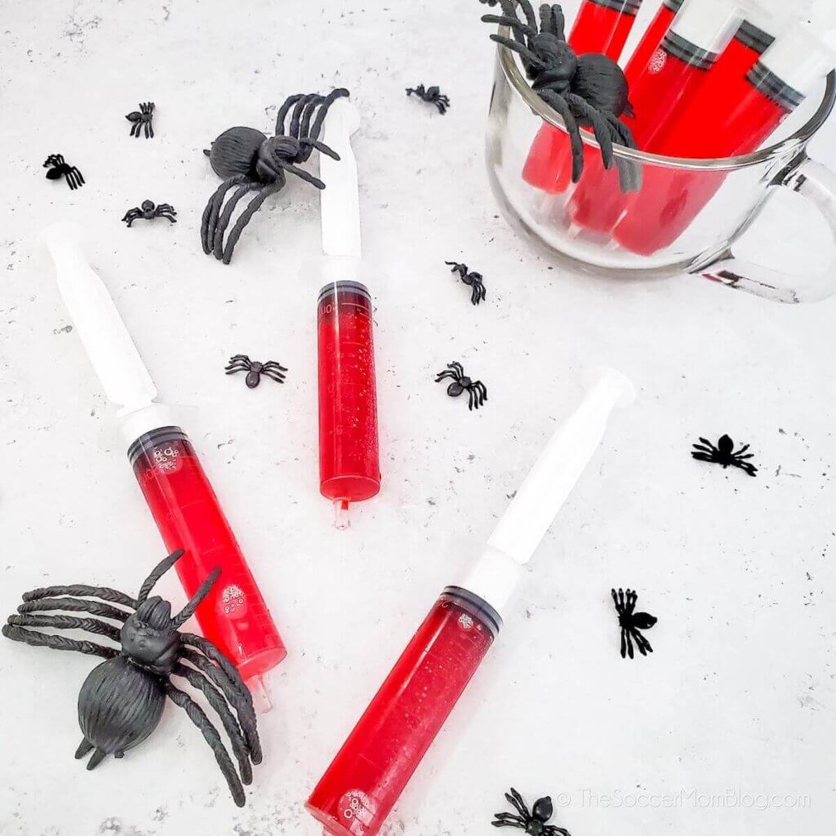 Red halloween jello shots in syringes surrounded by plastic spiders.