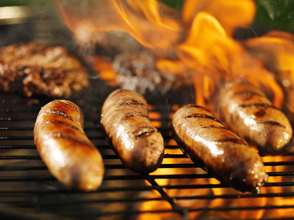 Brats cooking on a fiery grill with hamburgers.