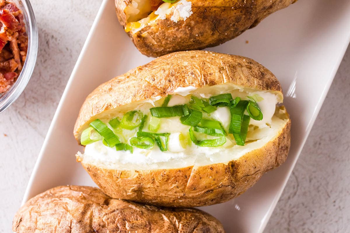 Baked potatoes on platter topped with butter, sour cream and thinly sliced green onions.
