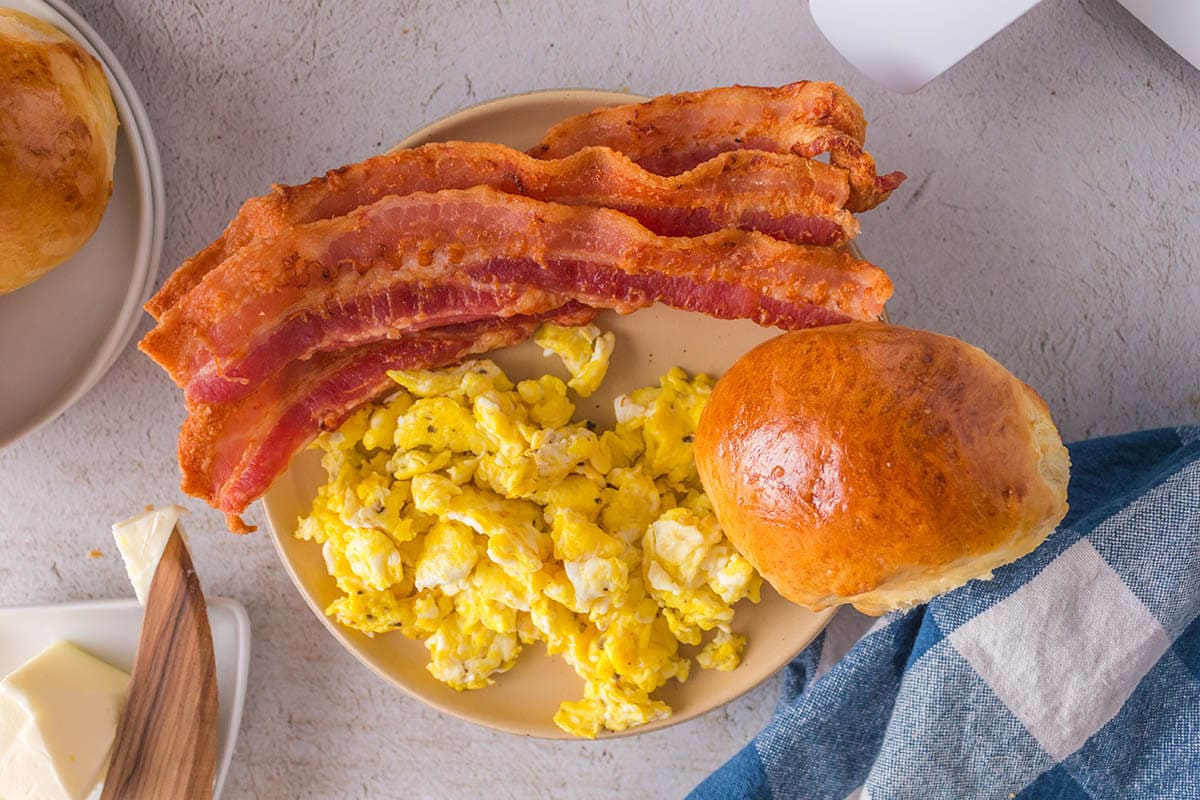 Plate filled with scrambled eggs, roll and crispy bacon.