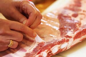Hands removing the membrane (silver skin) from the back of a raw rack of pork ribs before cooking.