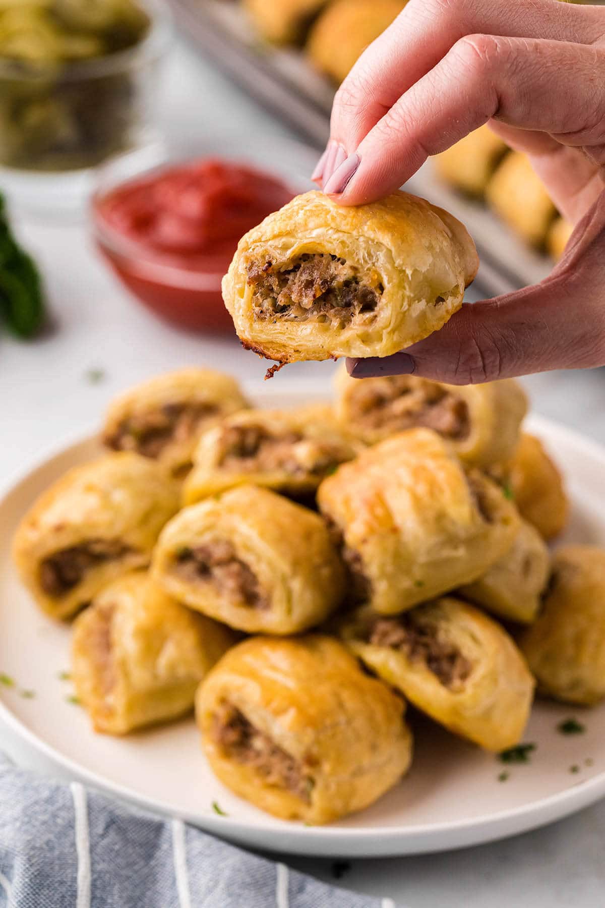 Puff Pastry Sausage Rolls on a platter with a hand holding one of the pastry rolls for a close up look.