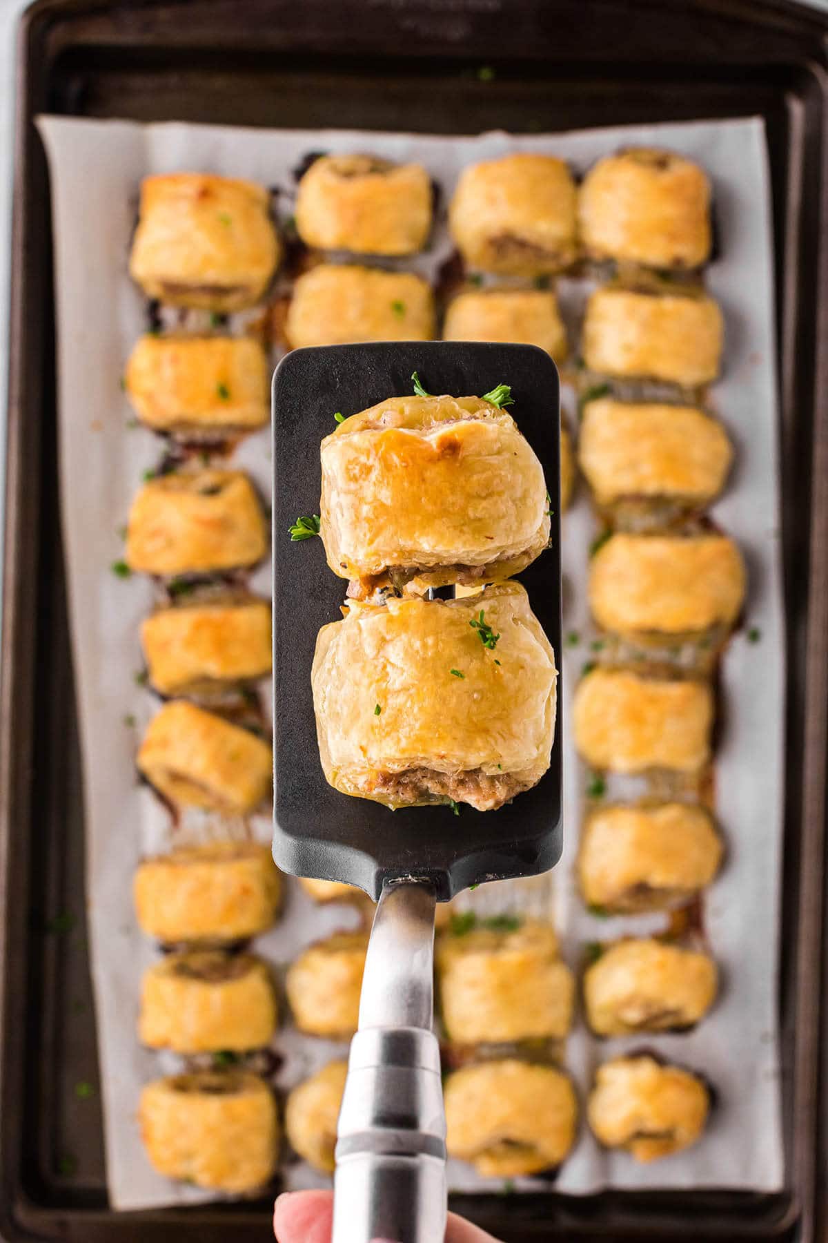 Puff pastry sausage rolls on a baking sheet with a spatula holding two of the rolls.