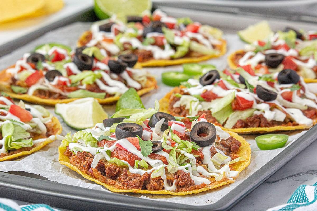 Sheet pan filled with ground beef tostadas. The tostadas are topped with salsa, lettuce, diced tomatoes, cilantro, and olives.