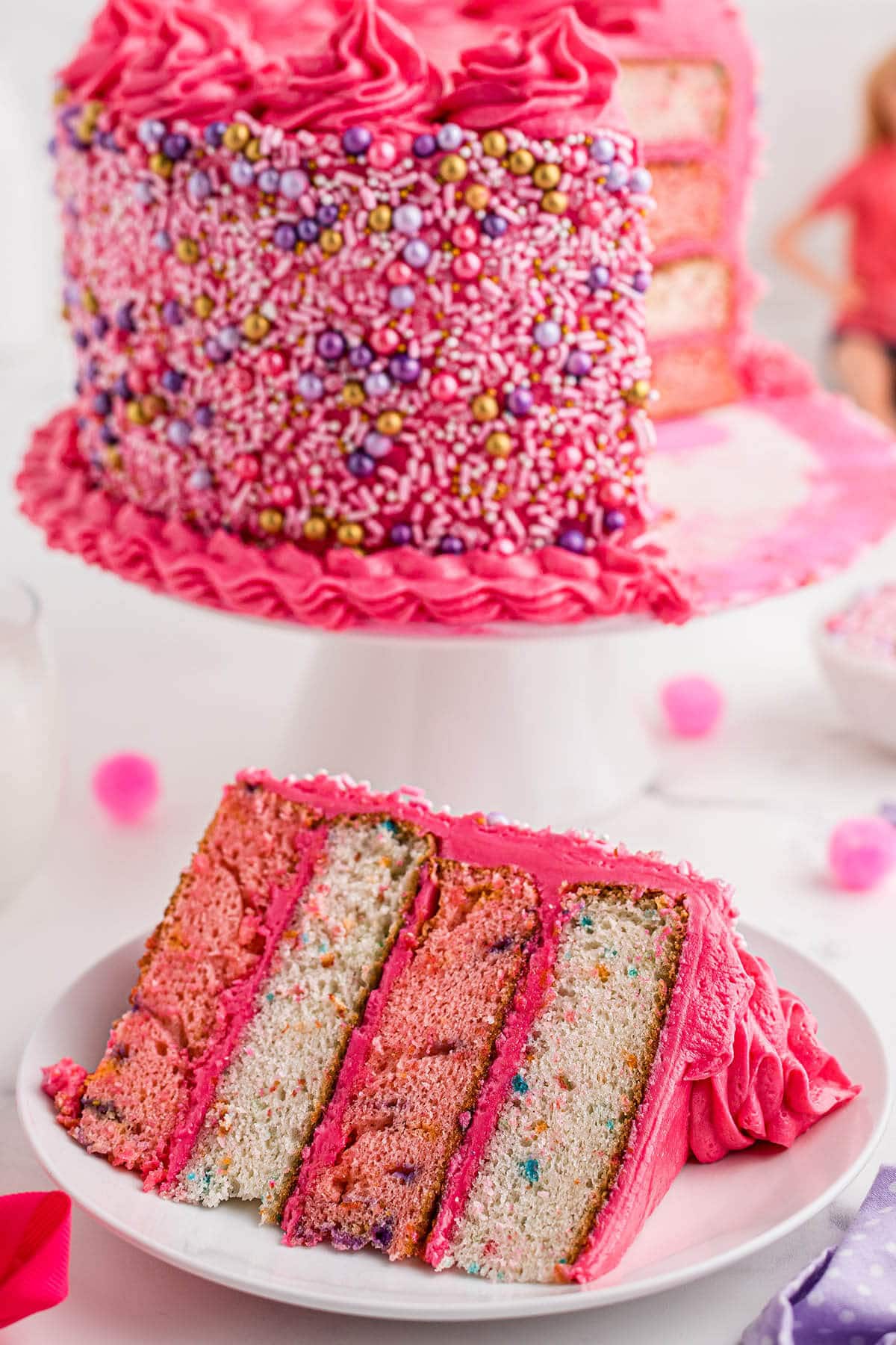 Slice of pink and white funfetti layer cake on a plate, with cake on a cakestand behind it.