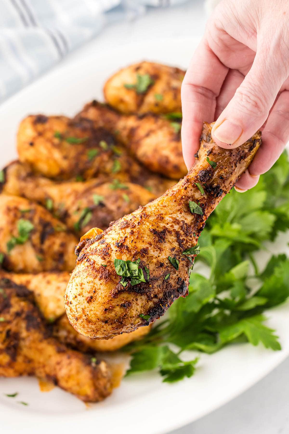 Grilled chicken drumsticks on a platter garnished with chopped parsley. A hand is holding a cooked chicken leg.