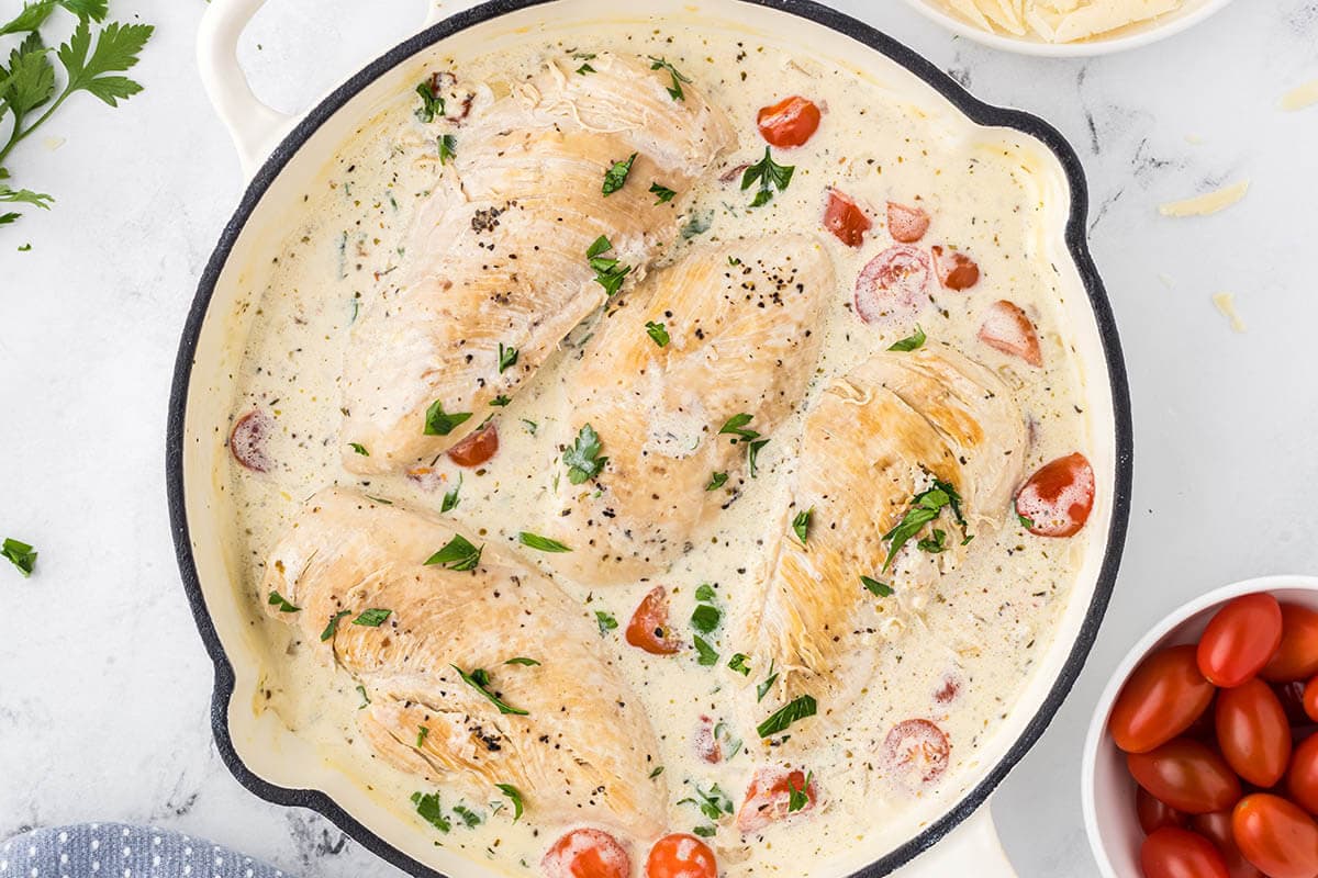 Chicken in a skillet being simmered in a creamy white sauce with cherry tomatoes.