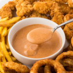 Red Robin Campfire Sauce in bowl with a serving spoon. It's surrounded by fries, chicken strips and onion rings.