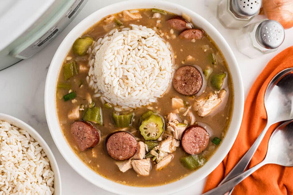 A bowl of gumbo with sliced sausage, okra, chicken pieces, and a serving of white rice on top. Silver spoons are beside the dish, along with a small bowl of rice and orange cloth. Salt and pepper shakers are in the background.