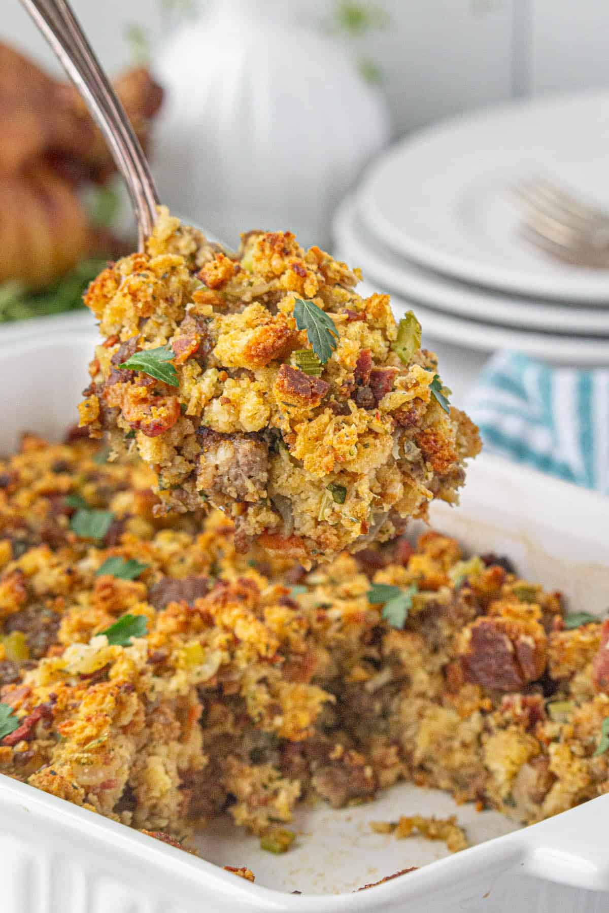 A casserole dish filled with sausage and bacon stuffing, with a serving spoon removing a big scoop.