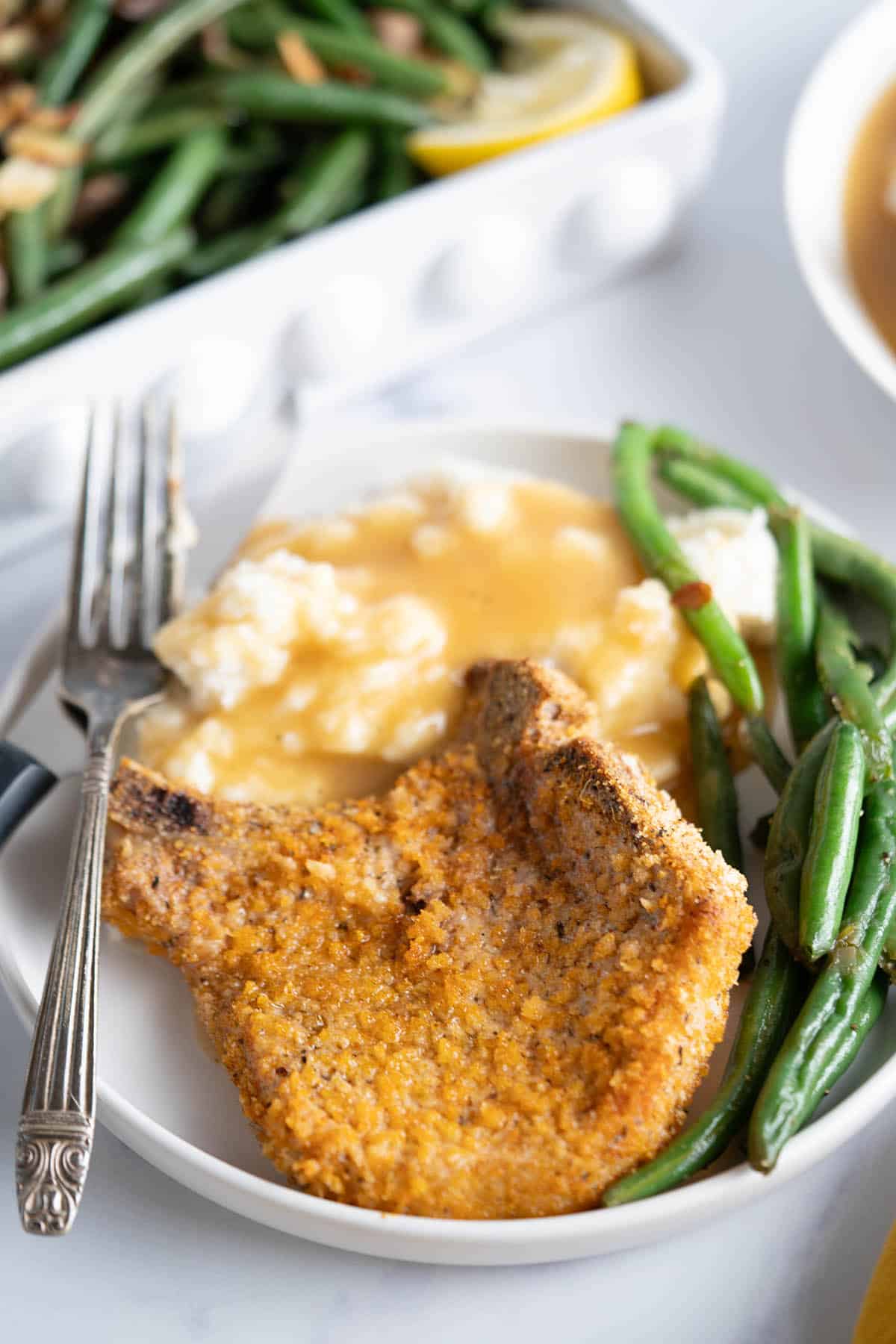 Crispy pork chop on a plate, served with green beans and mashed potatoes with gravy.