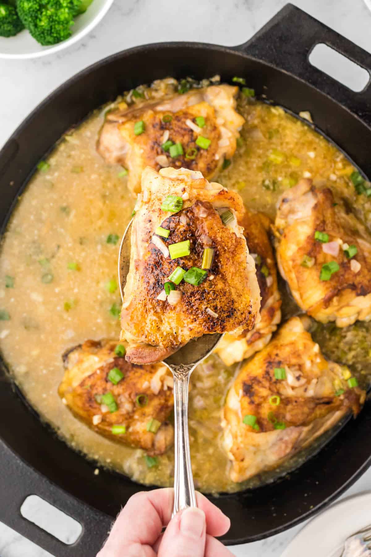Fried chicken thighs in a sauce, in a skillet, with a serving spoon.