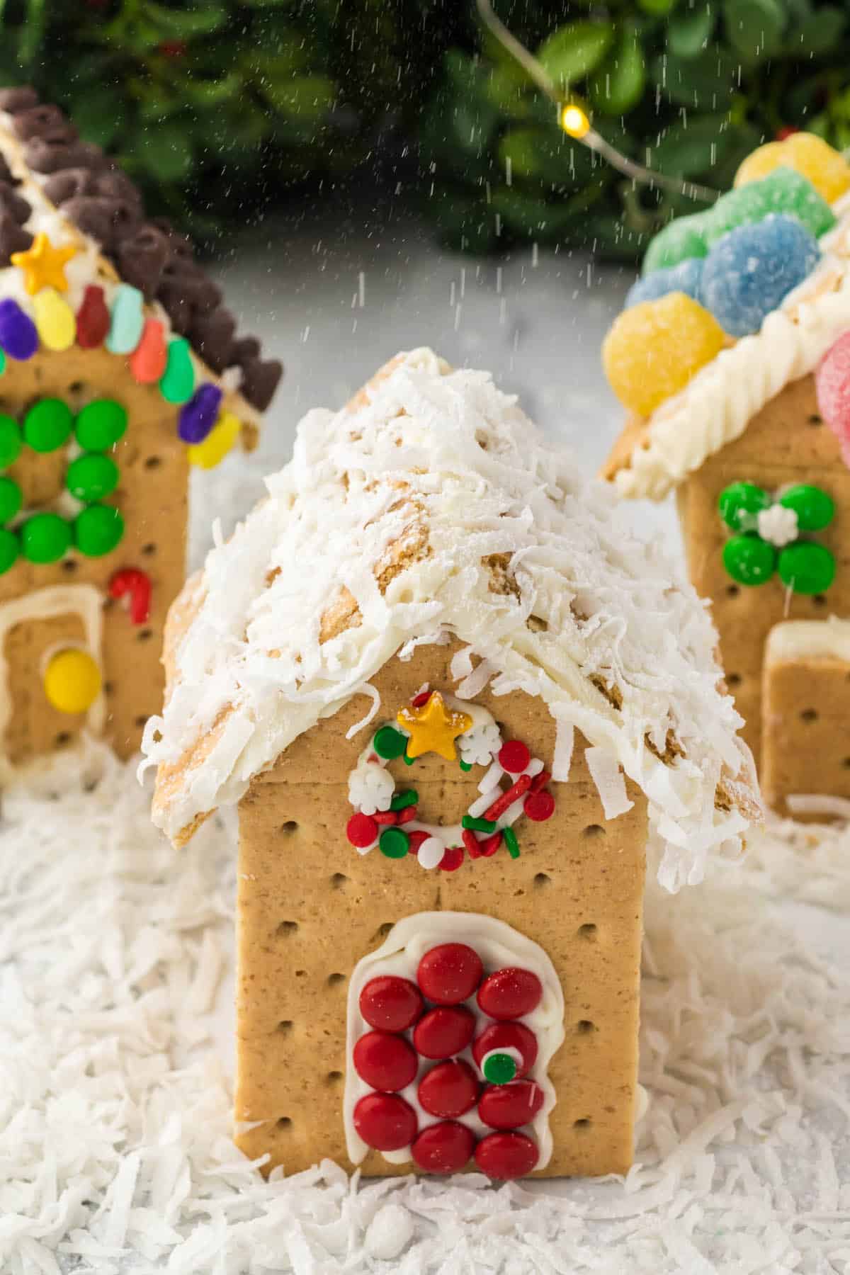 Three gingerbread houses on a bed of coconut snow with powdered sugar snow raining down on the candy houses.