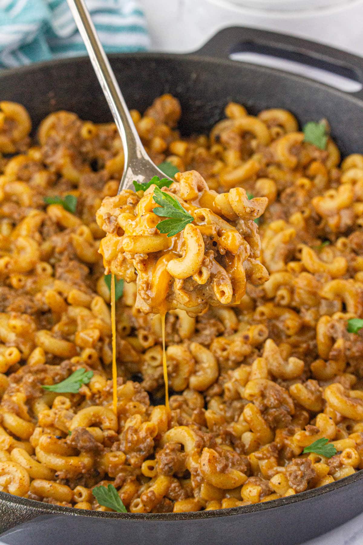 Cast iron skillet filled with homemade cheesy hamburger helper. With a serving spoon taking a scoop.