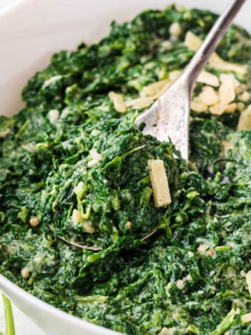 A large white bowl filled with Ruth Chris Steamed Spinach Recipe, with a serving spoon.