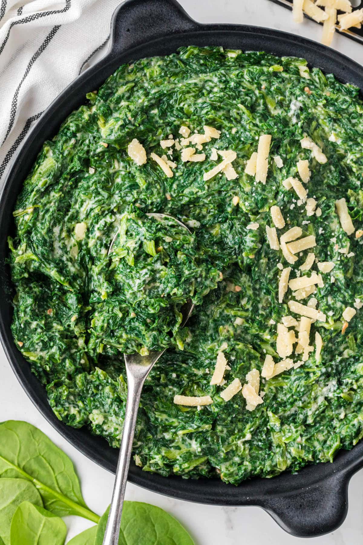 Skillet filled with creamed spinach topped with shaved parmesan cheese. With a serving spoon.
