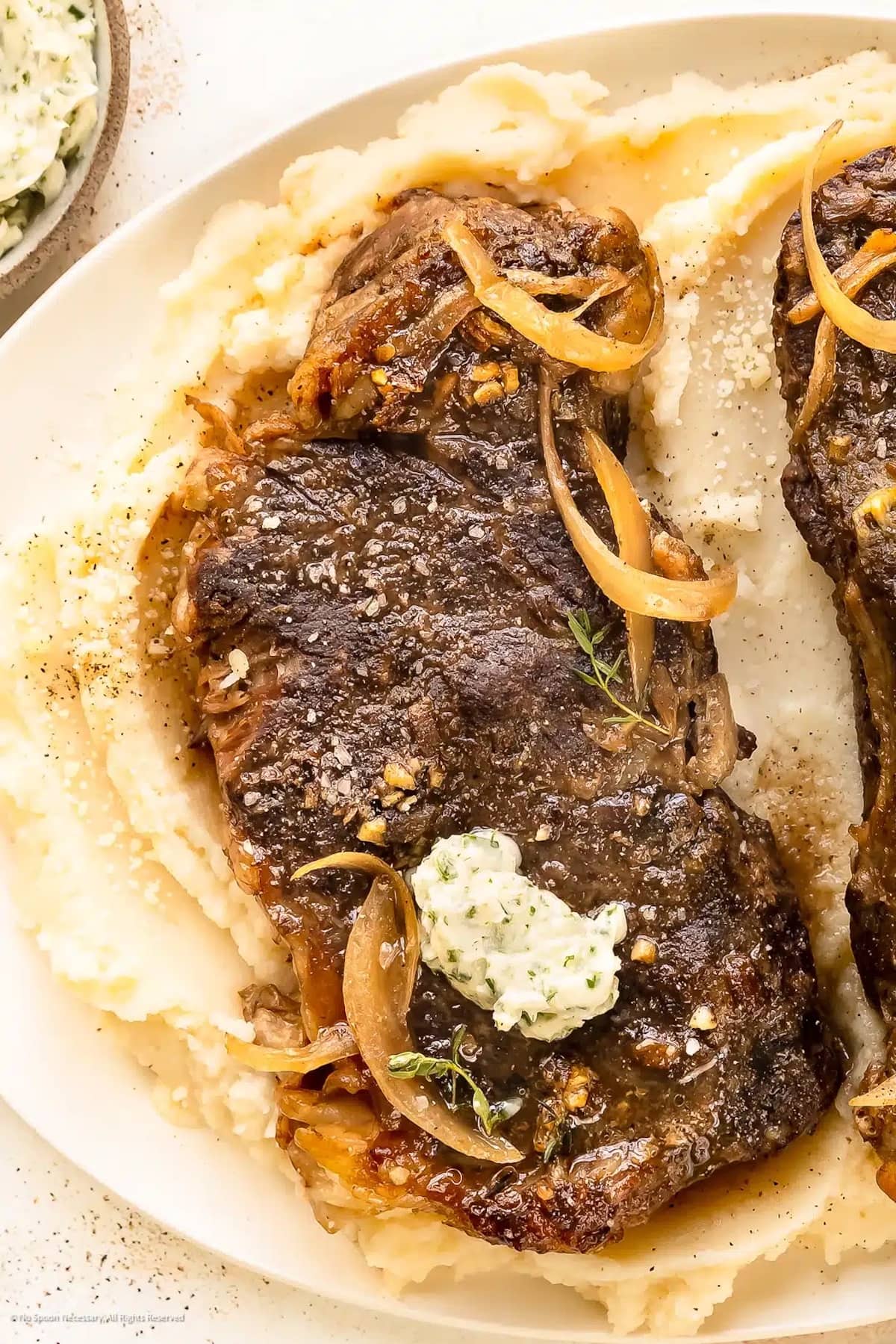 Ribeye Steak on a plate topped with butter served over a pile of mashed potatoes.
