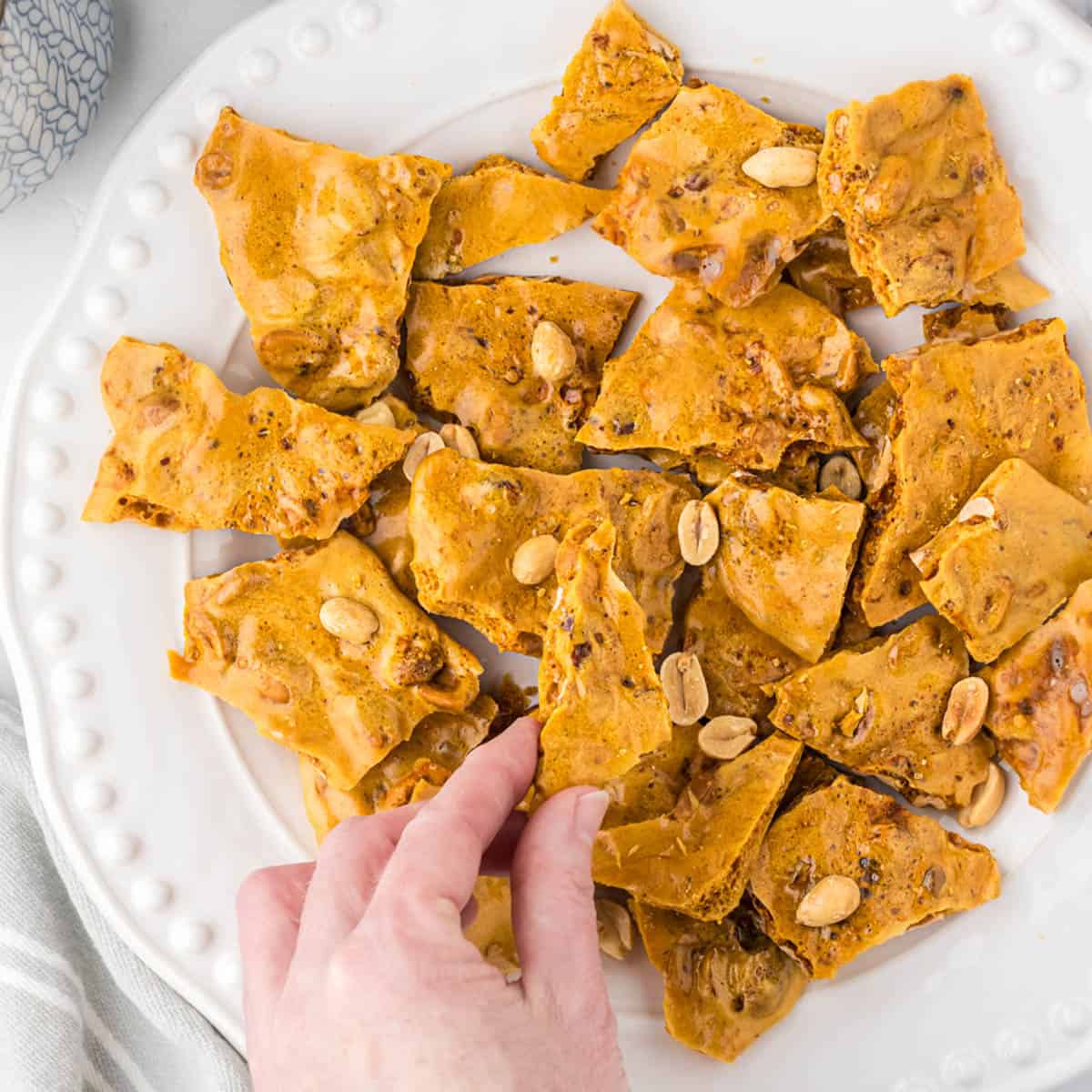 A platter of homemade peanut brittle and a hand is reaching in and grabbing a piece.