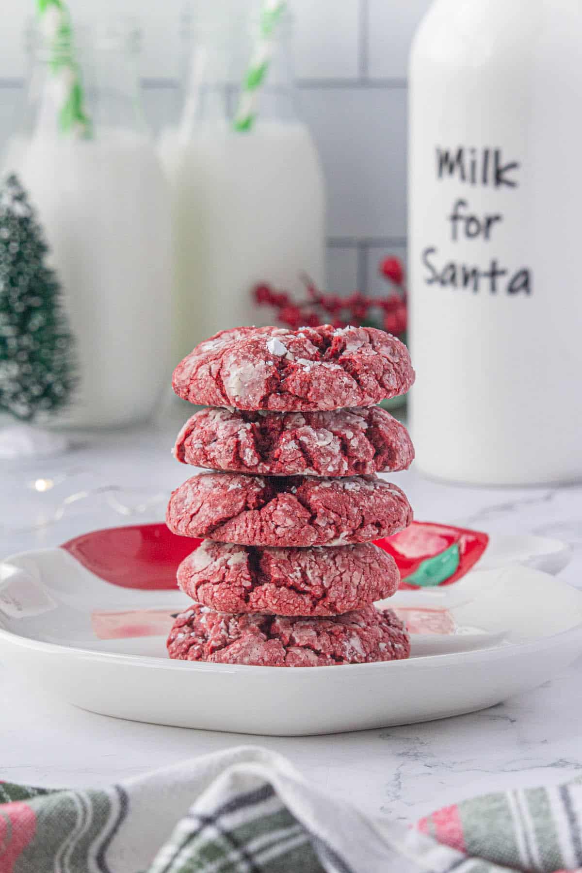 A stack of Red Velvet Crinkle Cookies on a holiday platter with glasses of milk for Santa in the background.