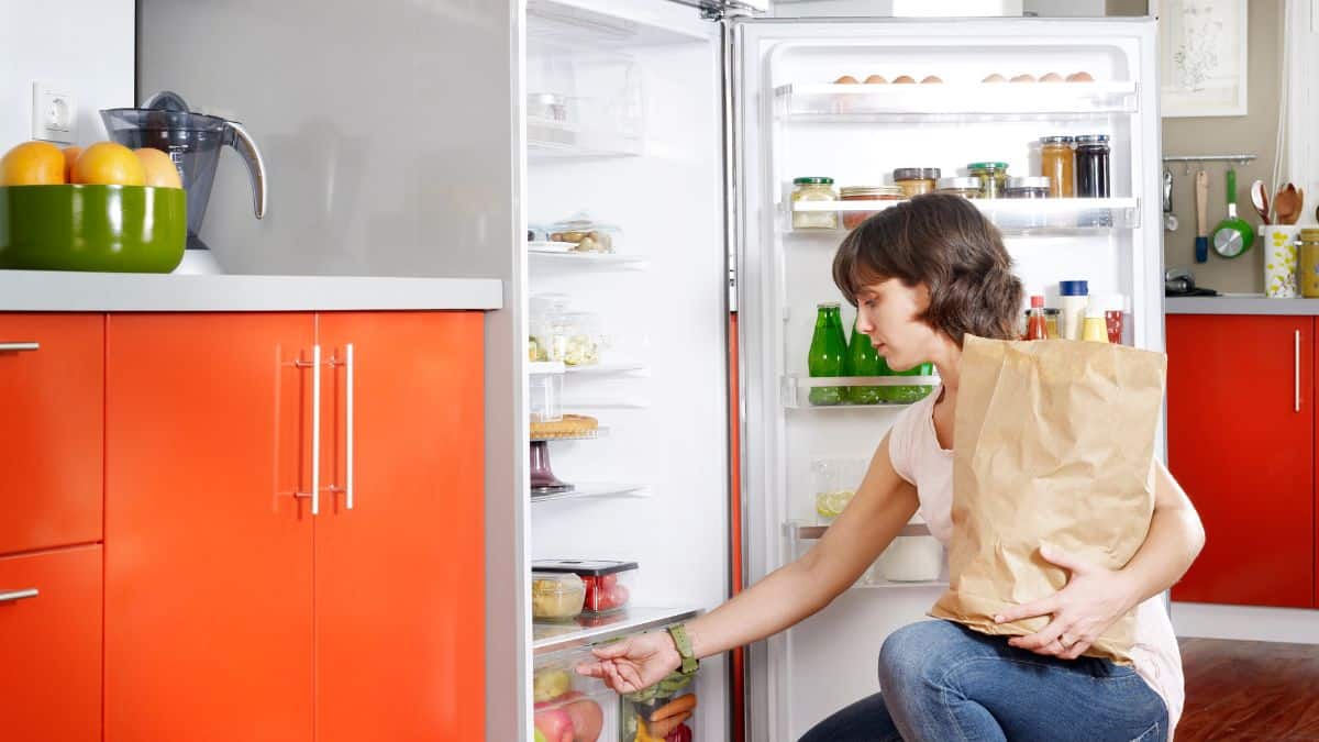 A woman kneeling in front of a refrigerator unloading groceries from a brown paper bag.