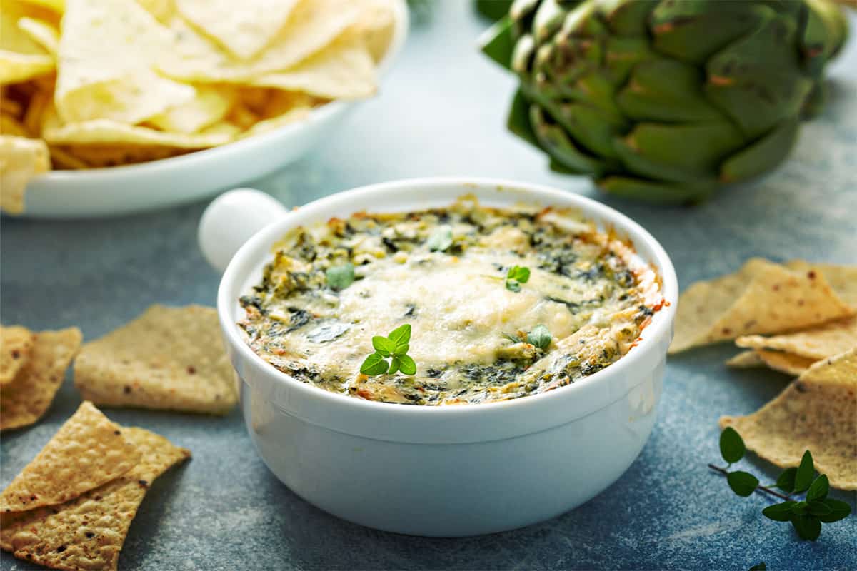 Baked spinach artichoke dip in a white bowl surrounded by tortilla chips.
