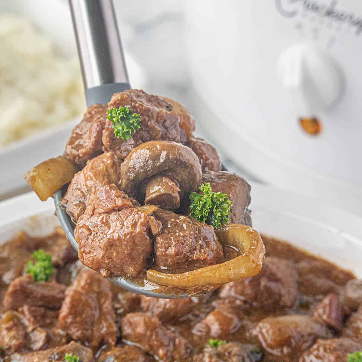 A spoon scooping up a hefty serving of crockpot steak bites in gravy with mushrooms and onions.