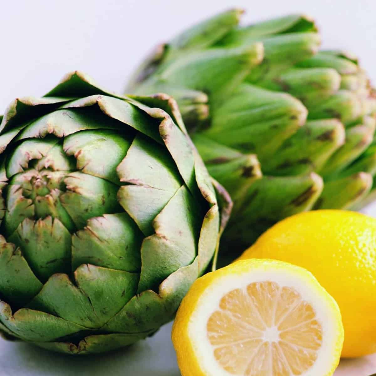 Fresh artichokes on a white counter, with sliced lemons on the side.