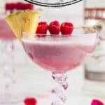 A Barbie Cocktail in a pretty glass garnished with fresh raspberries and a wedge of pineapple. With print overlay for Pinterest.