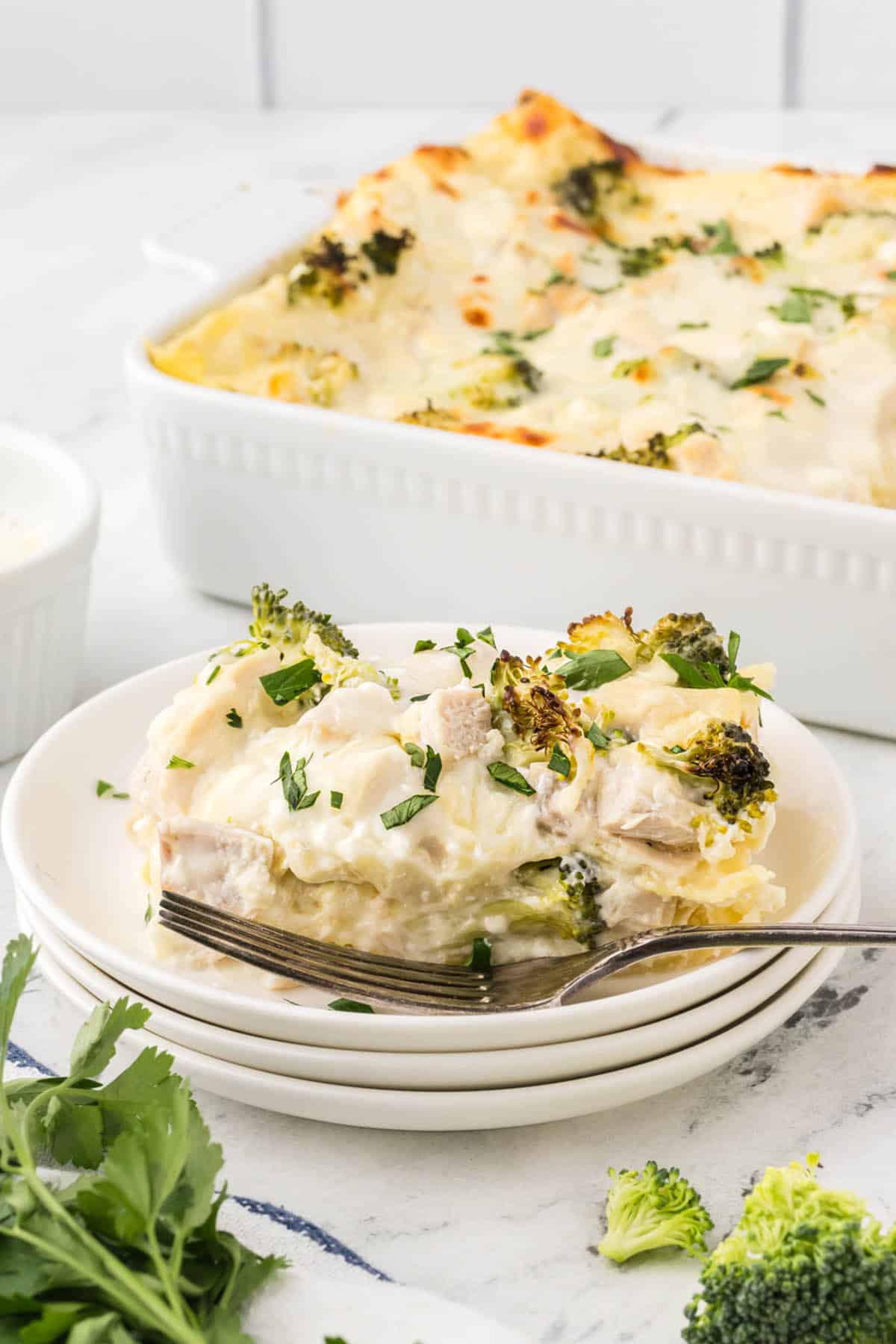 A slice of alfredo lasagna on a plate with a fork. The rest of the casserole is in the background.