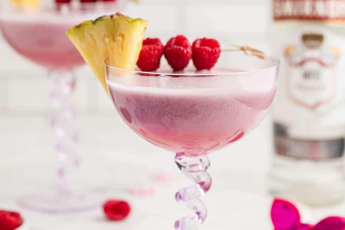 Pink Barbie Cocktail in a fun martini glass. Garnished with raspberries and pineapple.