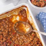A casserole dish filled with juicy bbq baked beans. A serving spoon is off to the side.