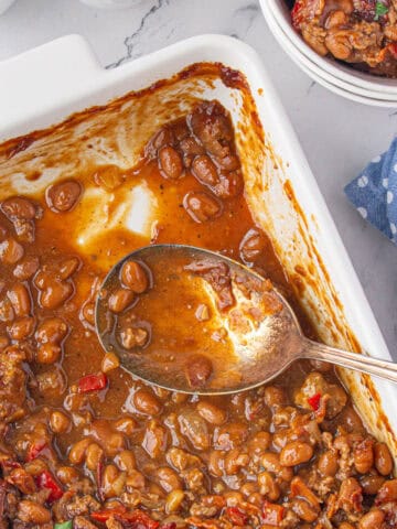 A casserole dish filled with juicy bbq baked beans. A serving spoon is off to the side.