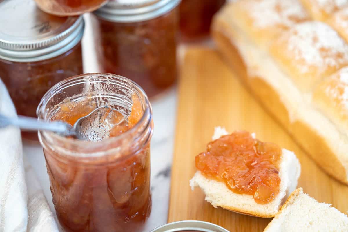 Pineapple Rhubarb jam in a jar with and rolls alongside topped with jelly.