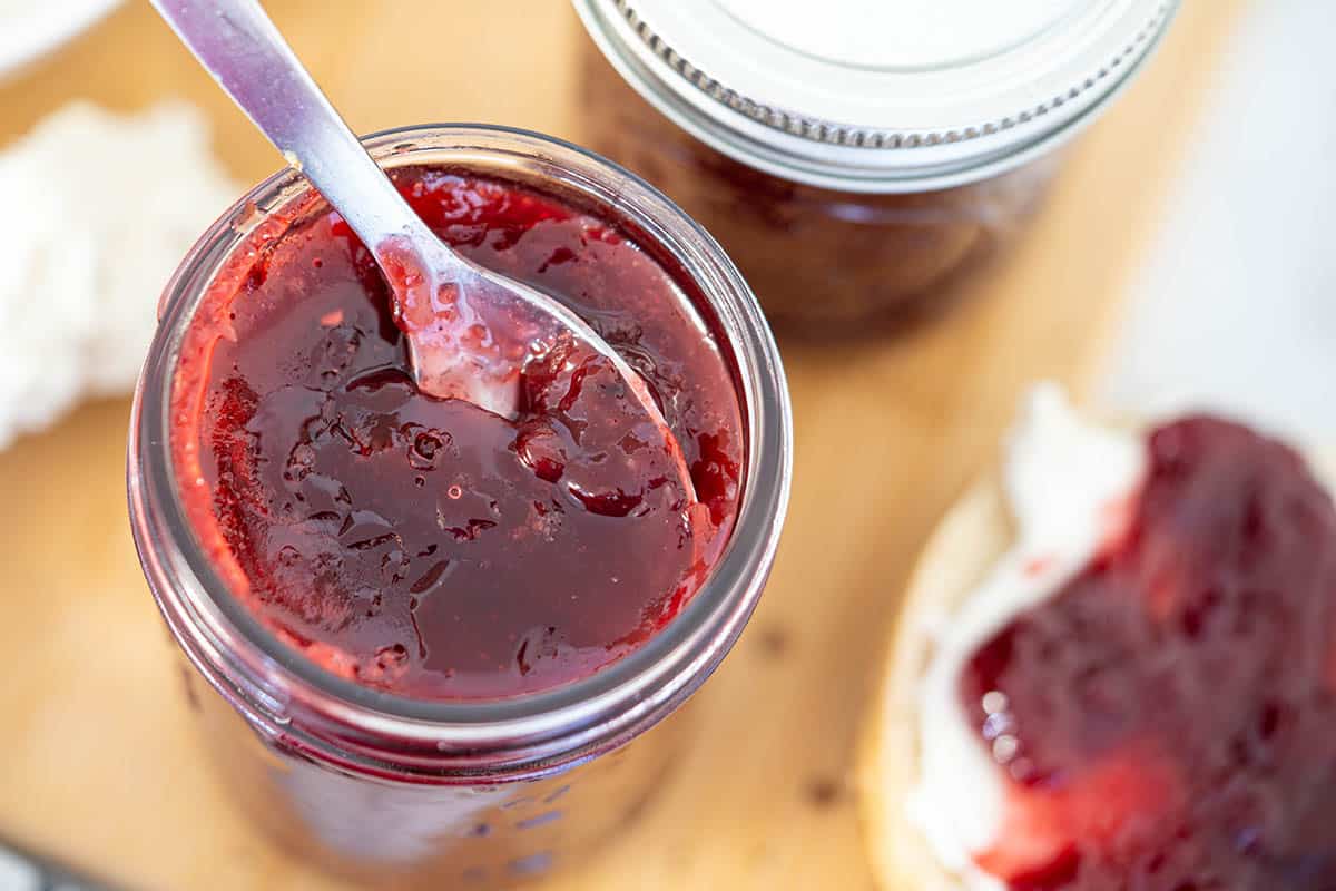 Jars of homemade cherry jam. On the side is a bagel topped with cream cheese and cherry jam.