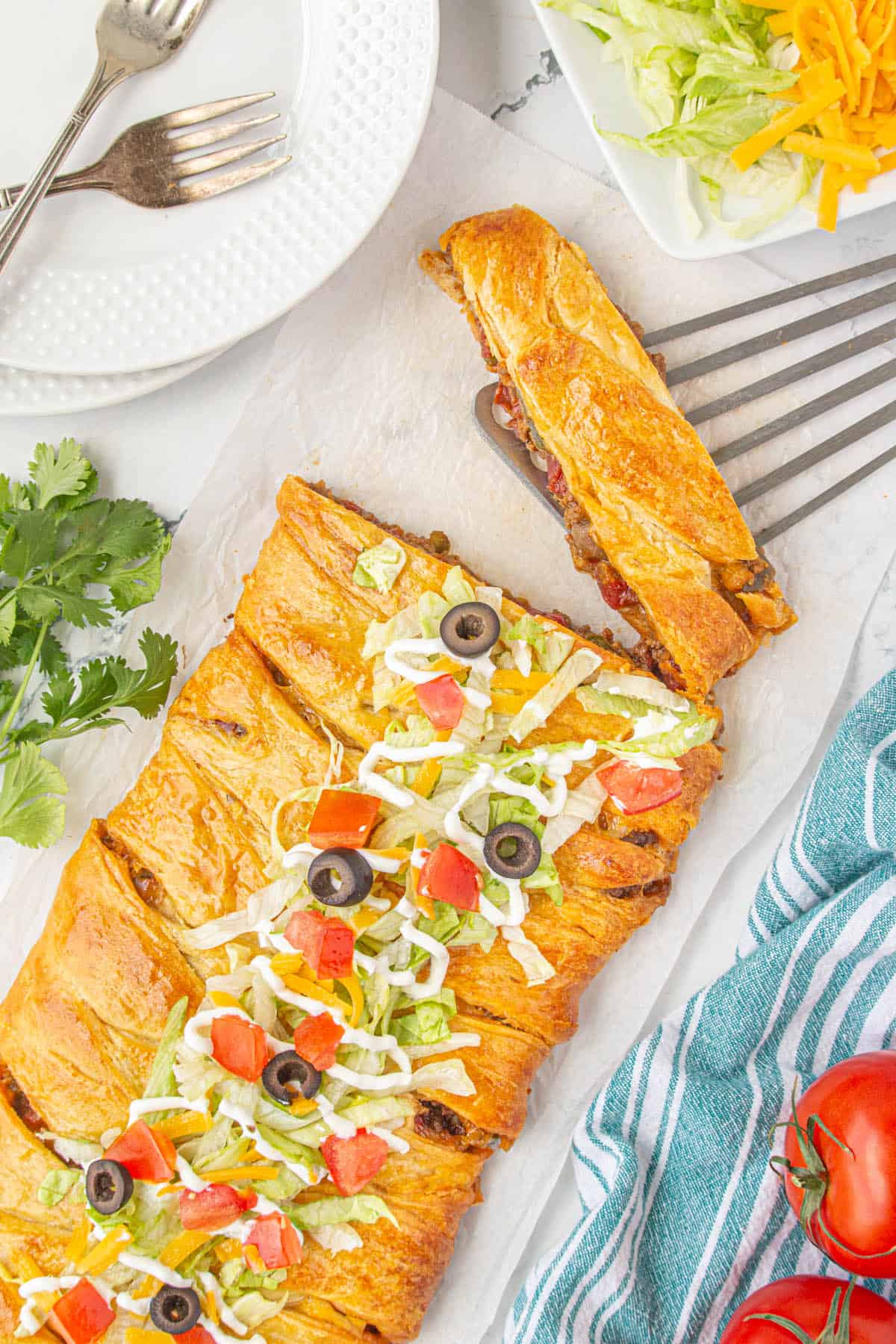 An overhead shot of fajita taco braid with a piece sliced off. The taco braid is baked until golden brown, topped with lettuce, sour cream, diced tomatoes and sliced olives.