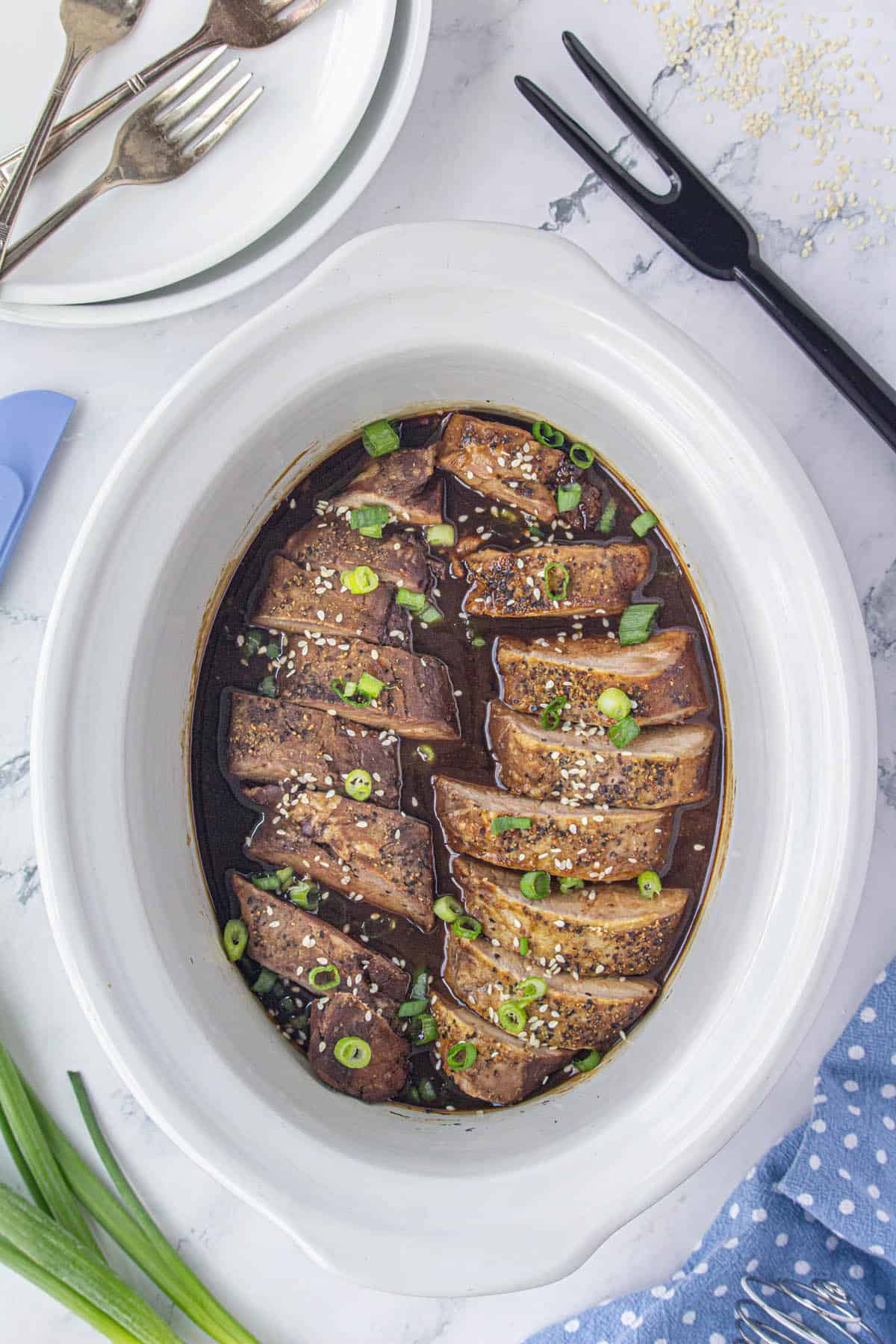 Sliced teriyaki pork loin in a crockpot garnished with sesame seeds and sliced onions.
