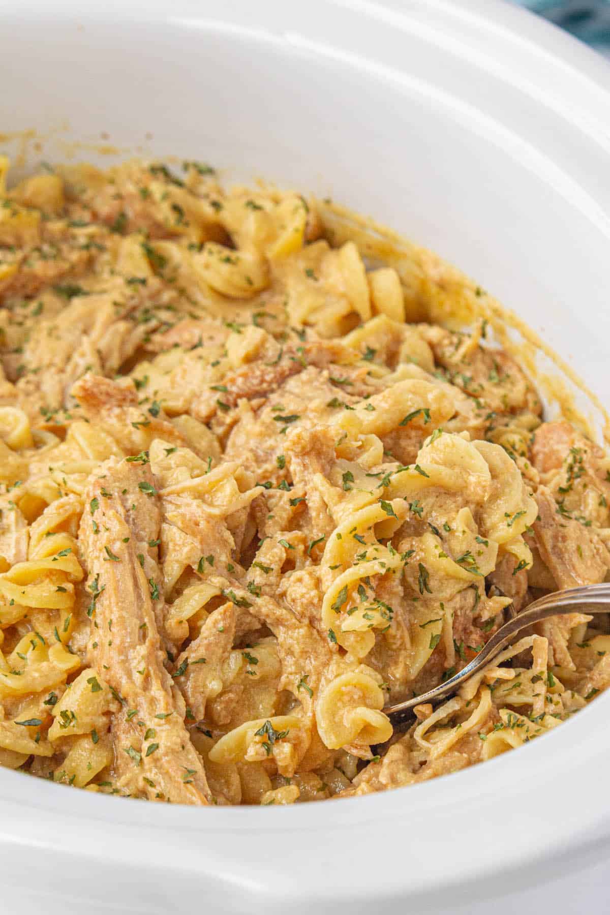 A slow cooker filled with chicken and noodles, with a spoon ready to take a scoop.