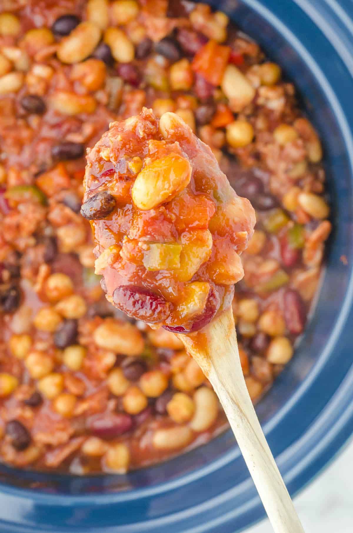 Loaded baked beans in a crockpot with a wood spoon grabbing a big scoop to serve them!