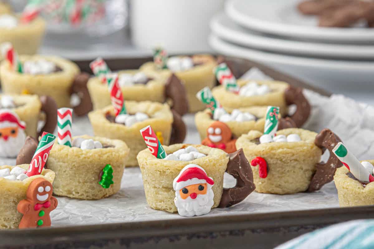 A cookie sheet filled with little cookies shaped to look like mugs of hot chocolate, decorated with candies on the outside.