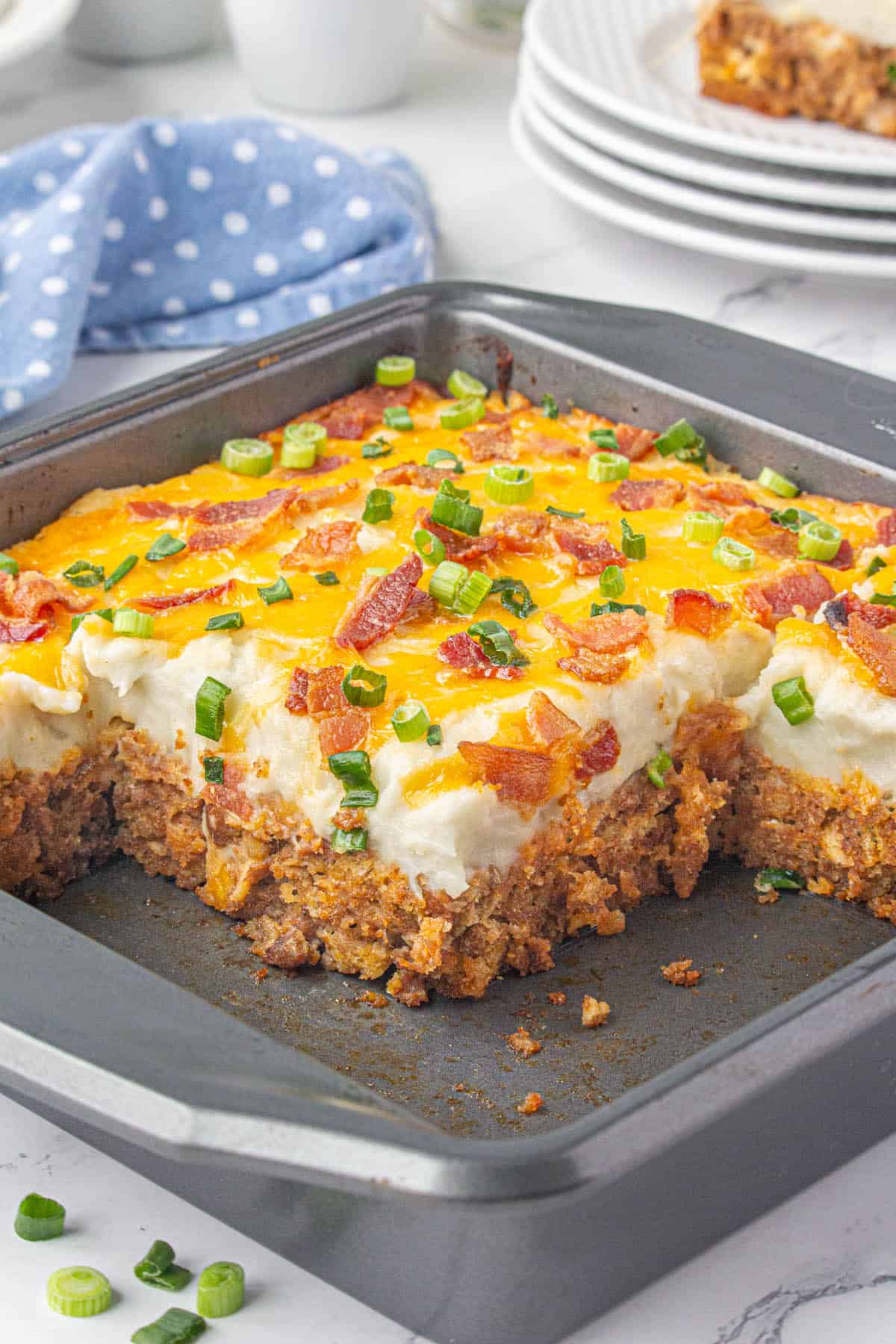 An 8" by 8" casserole dish filled with meatball potato casserole. With a couple of servings removed. The casserole is garnished with cheese, bacon and sliced green onions.