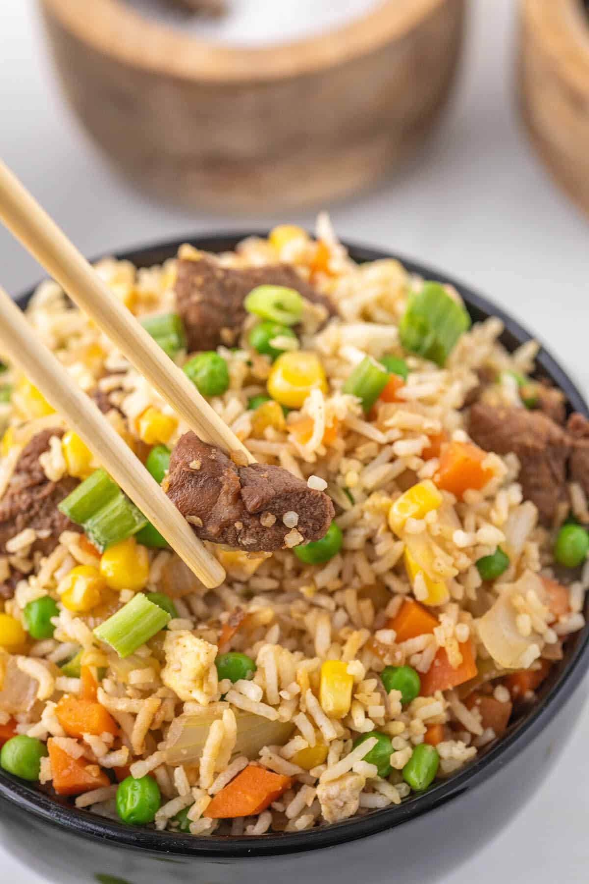 A black bowl filled with steak and fried rice, with a pair of chopsticks holding a piece of steak.