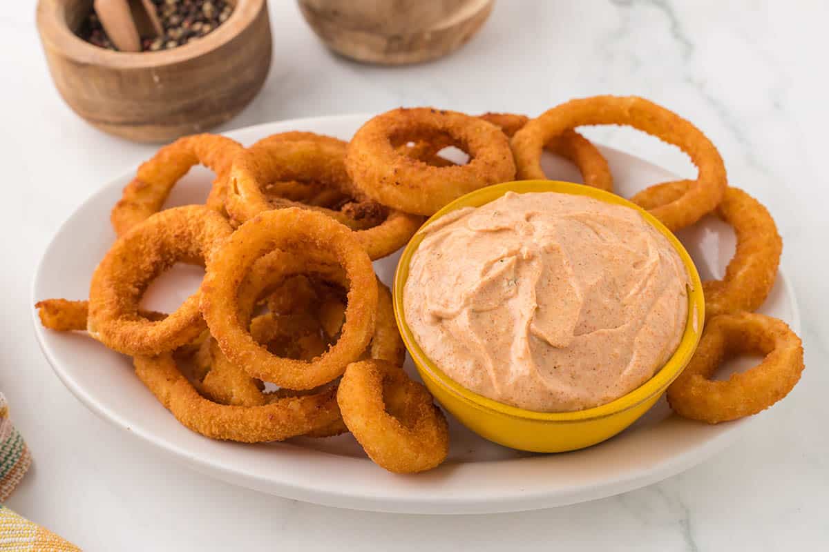 A platter of fried onion rings served with bowl filled with blooming onion sauce.