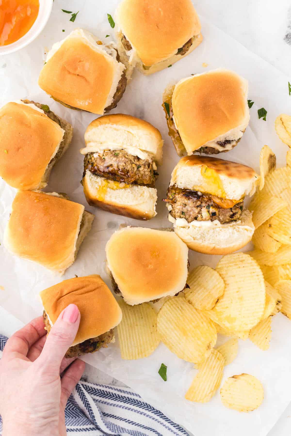 Mini burgers on a platter, served with chips. A hand is reaching in to grab a burger.