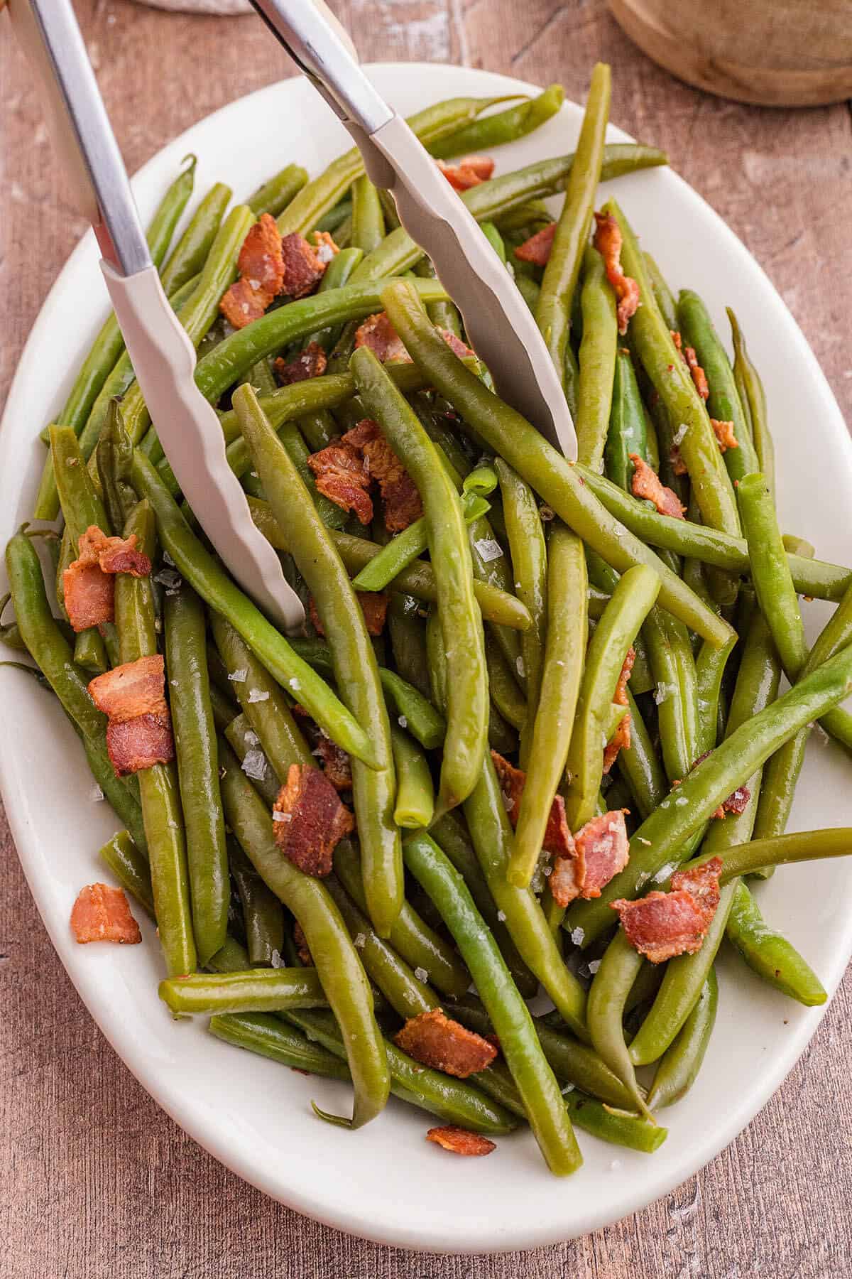 Green Beans topped with crispy bacon. A pair of tongs is dishing up a serving.