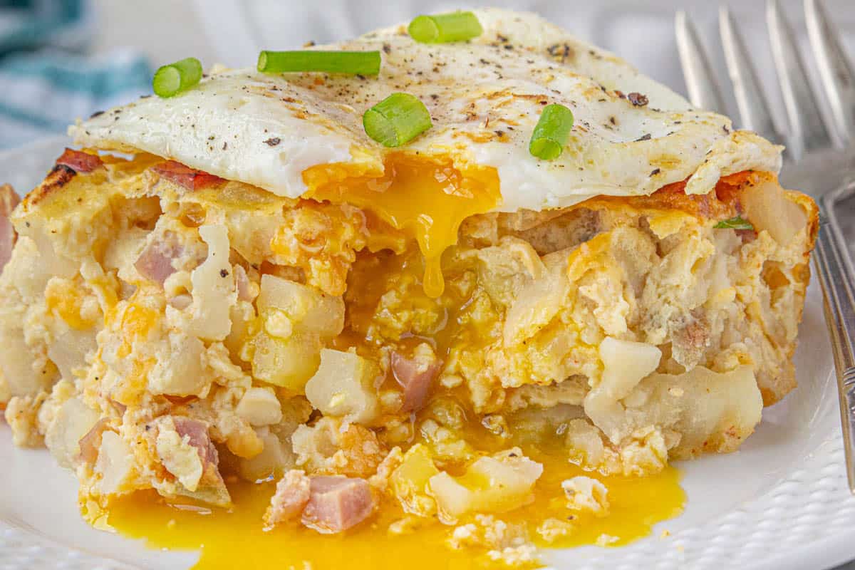 A slice of ham hashbrown breakfast casserole with a fried egg on top. The runny egg has been sliced into and is drizzling over the side of the slice.