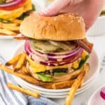 A steakhouse burger on a plate surrounded with fries. With a hand reaching in to grab a burger.