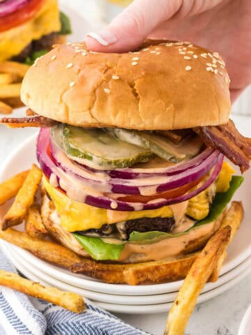 A steakhouse burger on a plate surrounded with fries. With a hand reaching in to grab a burger.